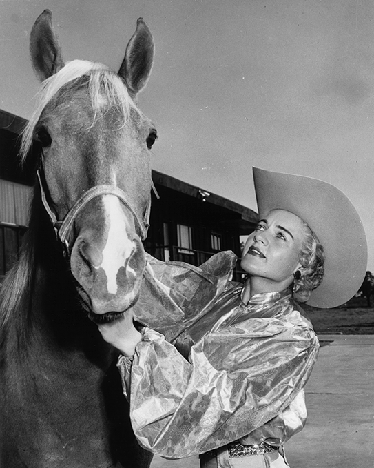 Woman wearing a wide-brimmed cowboy hat smiles while holding a horse by the bridle outdoors (black and white photo).