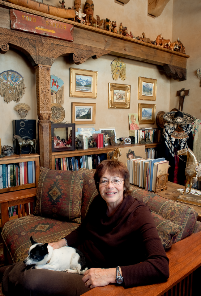 Smiling woman in a burgundy sweater sits on a patterned sofa with a small black-and-white dog on her lap in a cozy, cluttered living room.