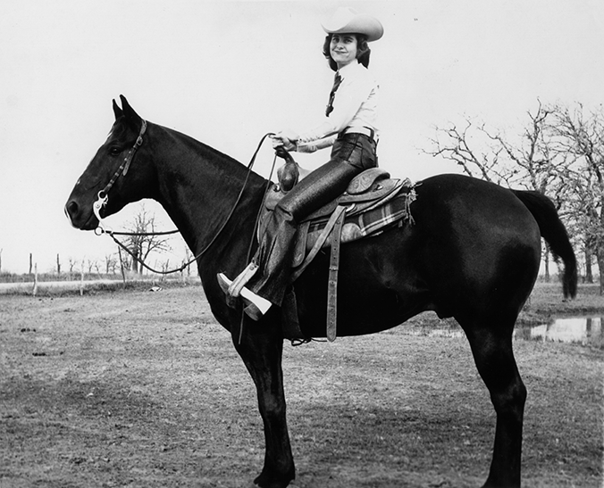 Woman in a cowboy hat riding a dark horse with a saddle in a rural, bare-field setting.