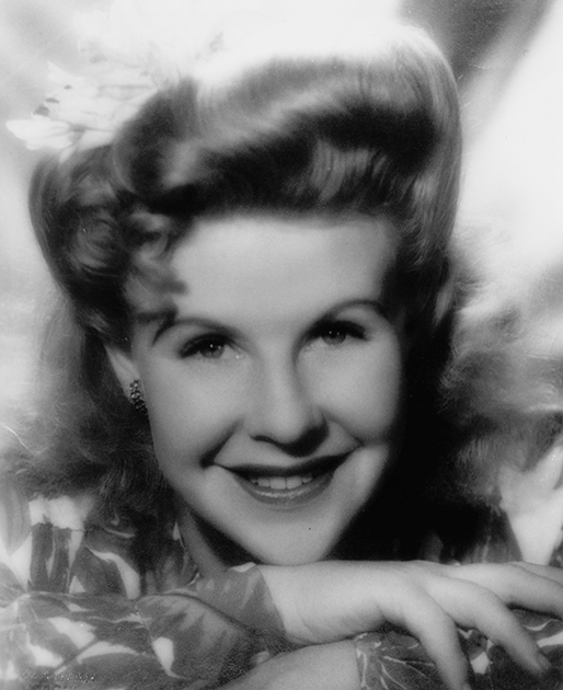 Black-and-white portrait of a smiling woman with waves in her hair, resting her folded arms on a surface.