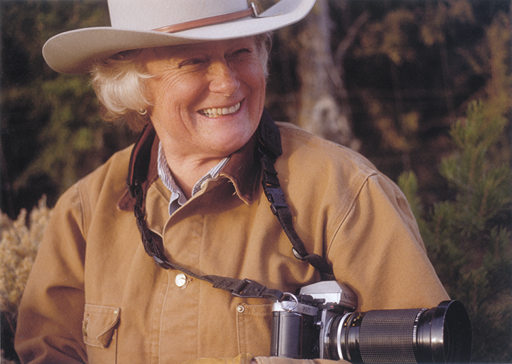 Smiling older woman in a cowboy hat with a camera hanging from her neck, outdoors in a natural setting.