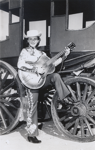 Cowgirl wearing a white hat and embroidered pants, playing an acoustic guitar beside a wooden-wheeled wagon.