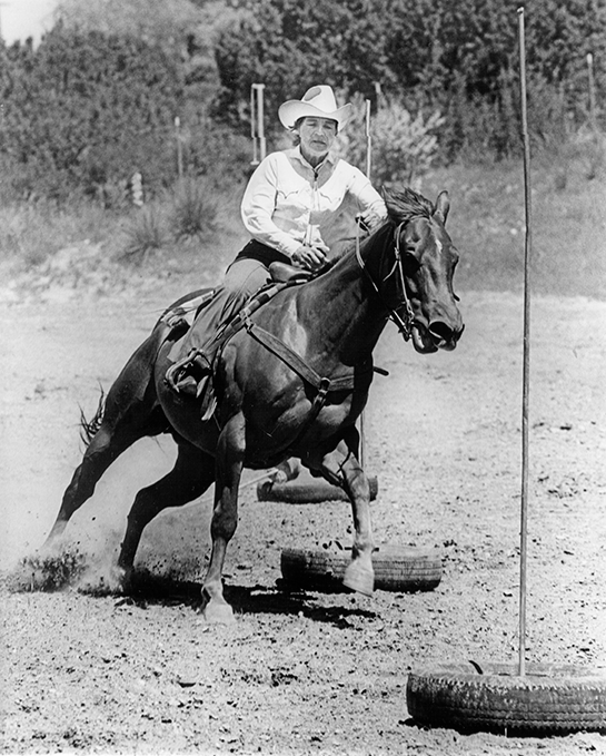 A cowboy in a white shirt and hat rides a horse through a dirt arena, navigating tires and vertical poles as obstacles.