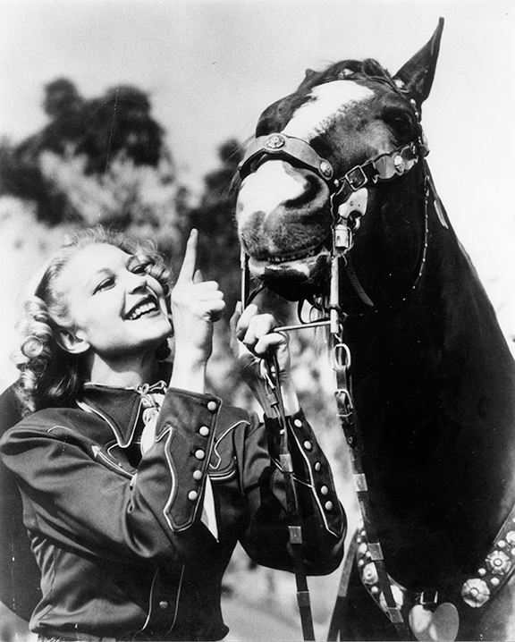 Cheerful woman in riding gear gesturing at a bridled horse during a sunny outdoor scene.