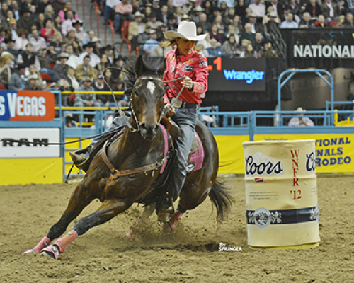 Cowgirl in a white hat and pink shirt rides a dark horse around a Coors-branded barrel in a crowded rodeo arena.