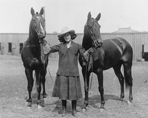 Woman wearing a wide-brim hat stands between two dark horses, each on a lead, in an outdoor paddock.