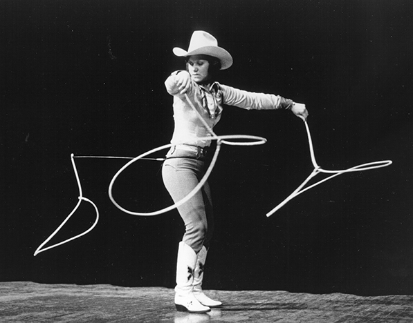 Cowboy in a hat and boots twirls a rope into looping lasso moves on a dark stage.