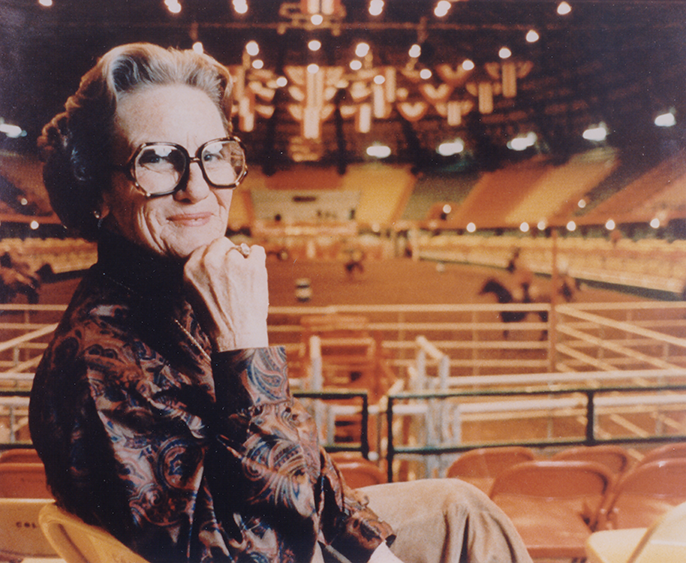 Older woman with large glasses sits in stadium seating, resting her chin on her hand and smiling, wearing a patterned blouse.