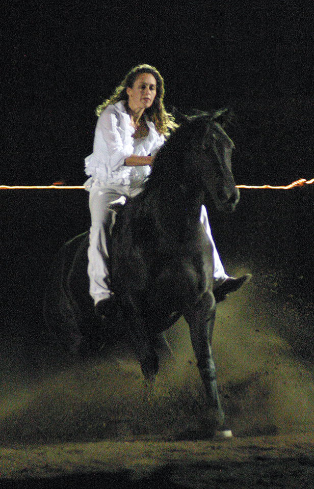 Woman in white riding a black horse at night, dust rising from hooves.