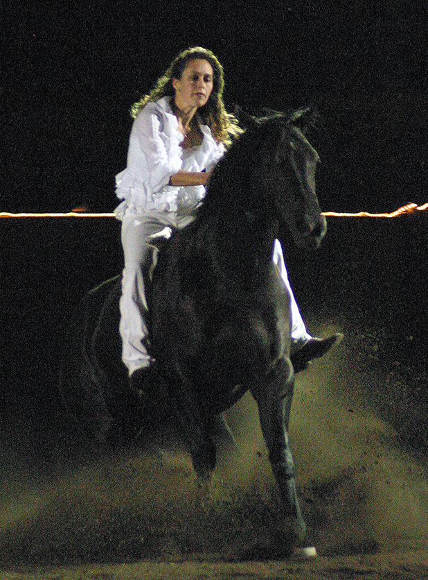 Woman in white riding a black horse at night, dust rising from hooves.