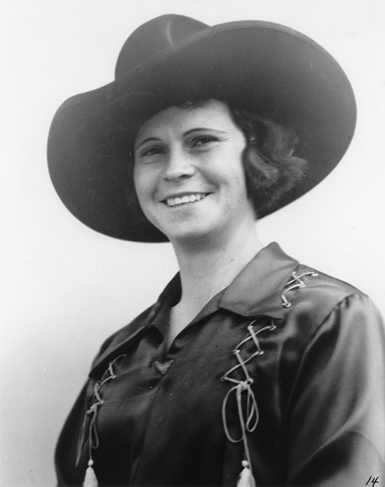 Smiling woman wearing a wide-brimmed hat and a western-style blouse with decorative lacing.