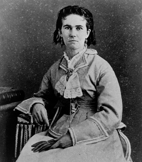Portrait of a woman in a Victorian-era dress seated with her hands on her lap, looking at the camera (black-and-white photo).