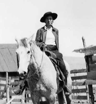 Person wearing a wide-brimmed hat rides a light-colored horse in a fenced corral on a ranch.