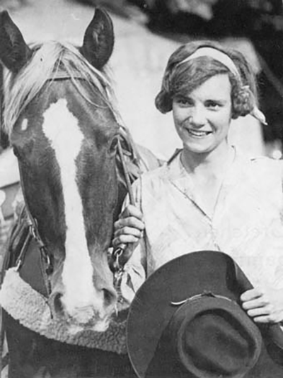Smiling woman in a light blouse standing beside a horse, holding a dark wide-brimmed hat.