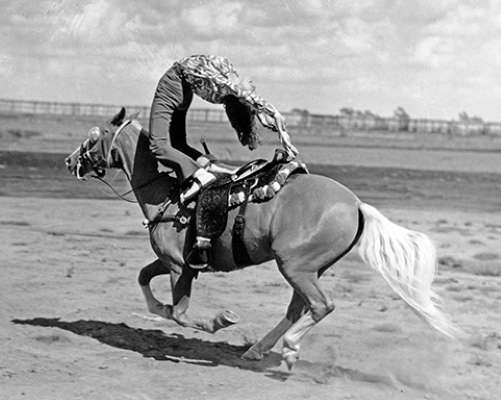 Black-and-white photo of a bareback rider on a bucking horse, dust rising as they race across a sandy beach.