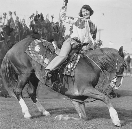 Woman riding a horse in a western-style outfit, raising her arm as the horse rears on a field (black-and-white vintage photo).