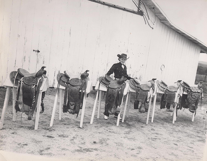 Black-and-white photo of a row of saddles on stands outside a wooden building, with a young person in a cowboy hat standing among them.