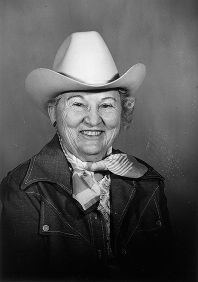 Smiling older woman wearing a white cowboy hat, scarf, and denim jacket in a black-and-white portrait.