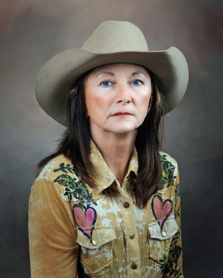 Woman wearing a beige cowboy hat and a tan floral shirt with heart patches, against a neutral backdrop (portrait).