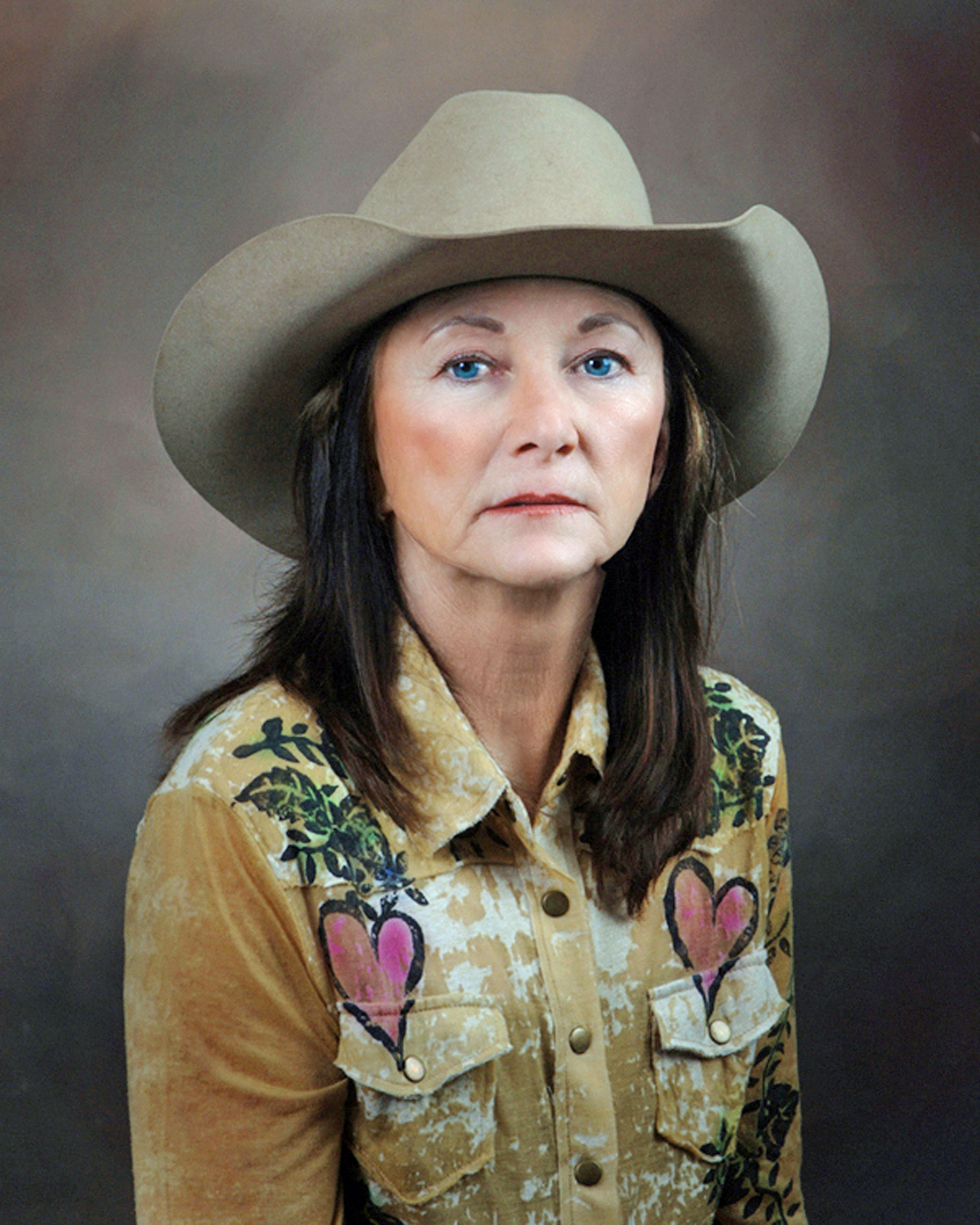 Woman wearing a beige cowboy hat and a tan floral shirt with heart patches, against a neutral backdrop (portrait).