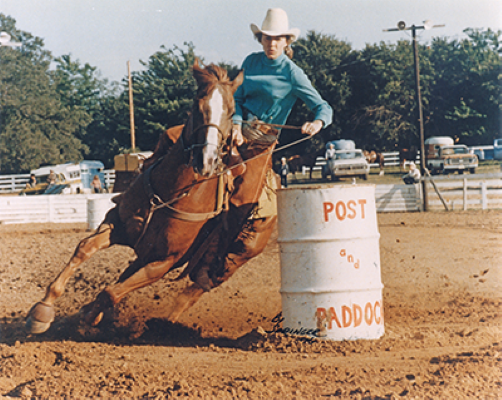 Rider in a turquoise shirt and white hat guides a horse around a white barrel in a dusty rodeo arena (barrel racing).