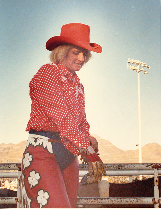Person wearing a red cowboy hat, red polka-dot shirt, and floral chaps leaning on a railing at a rodeo arena.
