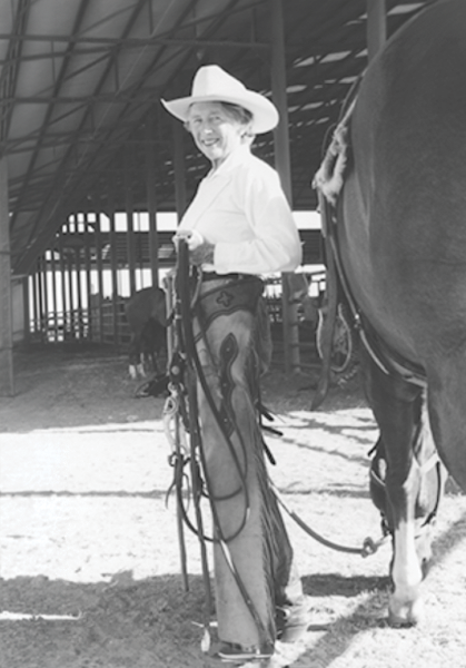 Older woman in a cowboy hat standing beside a horse in a barn, holding the reins.