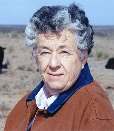 Portrait of an older woman with short gray hair, wearing a brown jacket, in a dry desert landscape with a blue sky behind her.