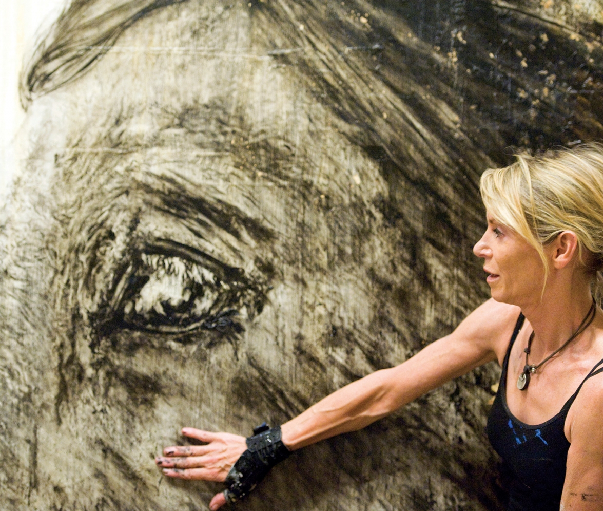 Woman in a black tank top touches a large grayscale mural of a horse's eye on a textured wall.