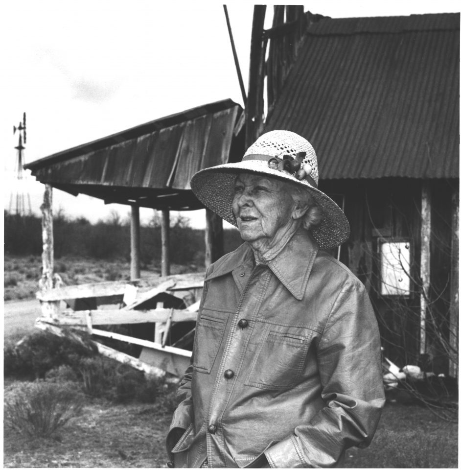 Older woman wearing a wide-brimmed hat and buttoned jacket standing outdoors near a rustic building.
