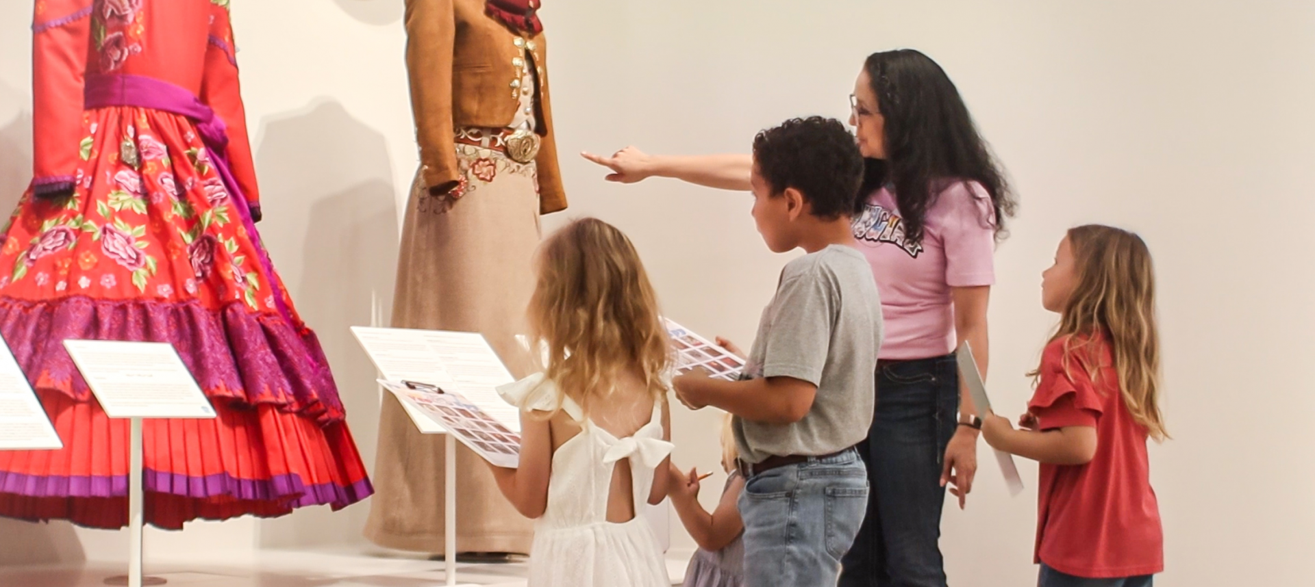 Children examine mannequined dresses on display with informational placards nearby in a museum setting.