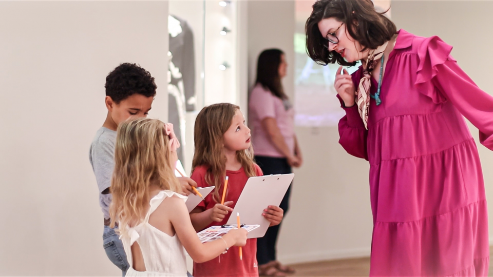 Teacher in a bright pink dress leaning toward four children holding clipboards and pencils, engaging in a learning activity in a classroom or gallery.