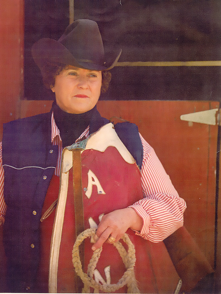 Older woman in a cowboy hat and navy vest stands holding a red book with white letters and a rope lasso in front of a wooden background, a portrait of western attire.