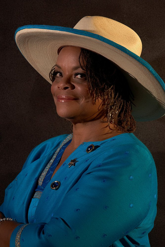 Portrait of a smiling woman in a turquoise jacket and a wide-brim straw hat with a blue ribbon against a dark background.