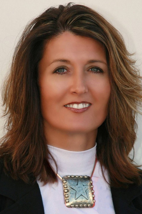 Portrait of a smiling woman with shoulder-length brown hair, wearing a white top, black blazer, and a large square silver star pendant necklace.