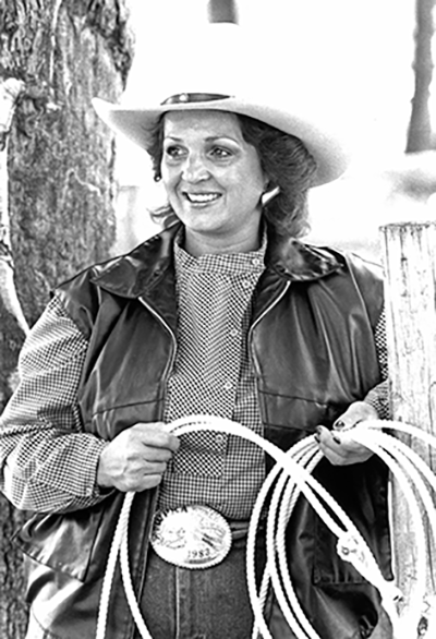 Smiling woman in a cowboy hat and leather vest coils a rope outdoors.