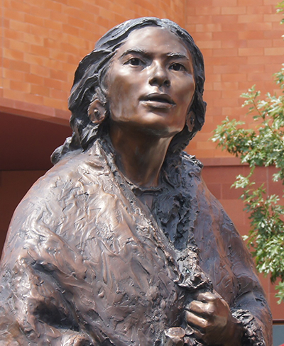 Bronze statue of a Native American woman in a shawl, outdoors beside a red brick building.