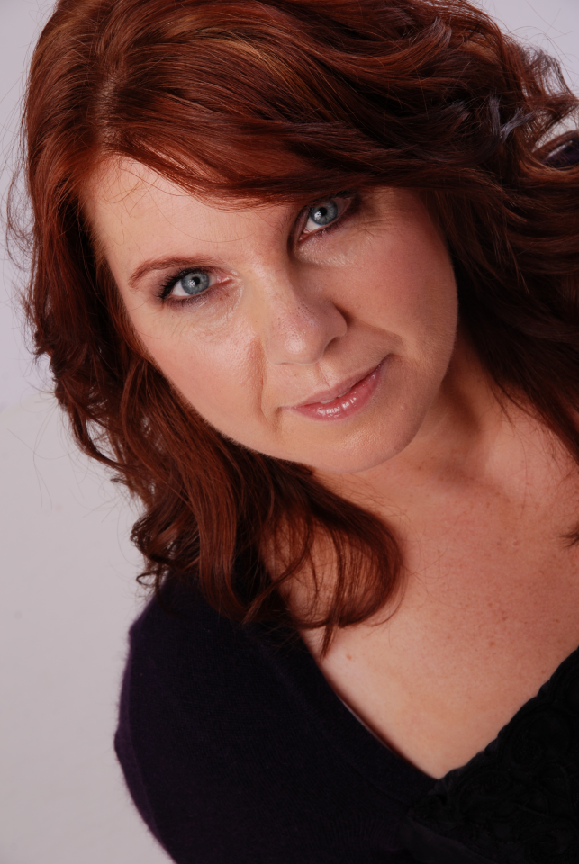 Close-up portrait of a smiling woman with auburn hair and blue eyes, wearing a dark top.