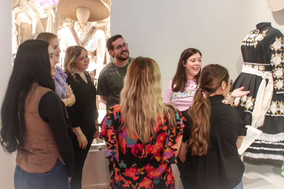Group of people standing and chatting near a large floral dress on a mannequin in a museum or shop display, with paper brochures in hand.