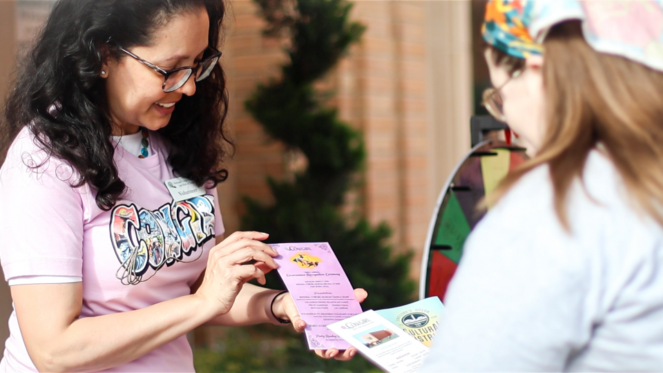 Woman in pink shirt hands pamphlets to someone wearing a light jacket outside, smiling and wearing glasses.