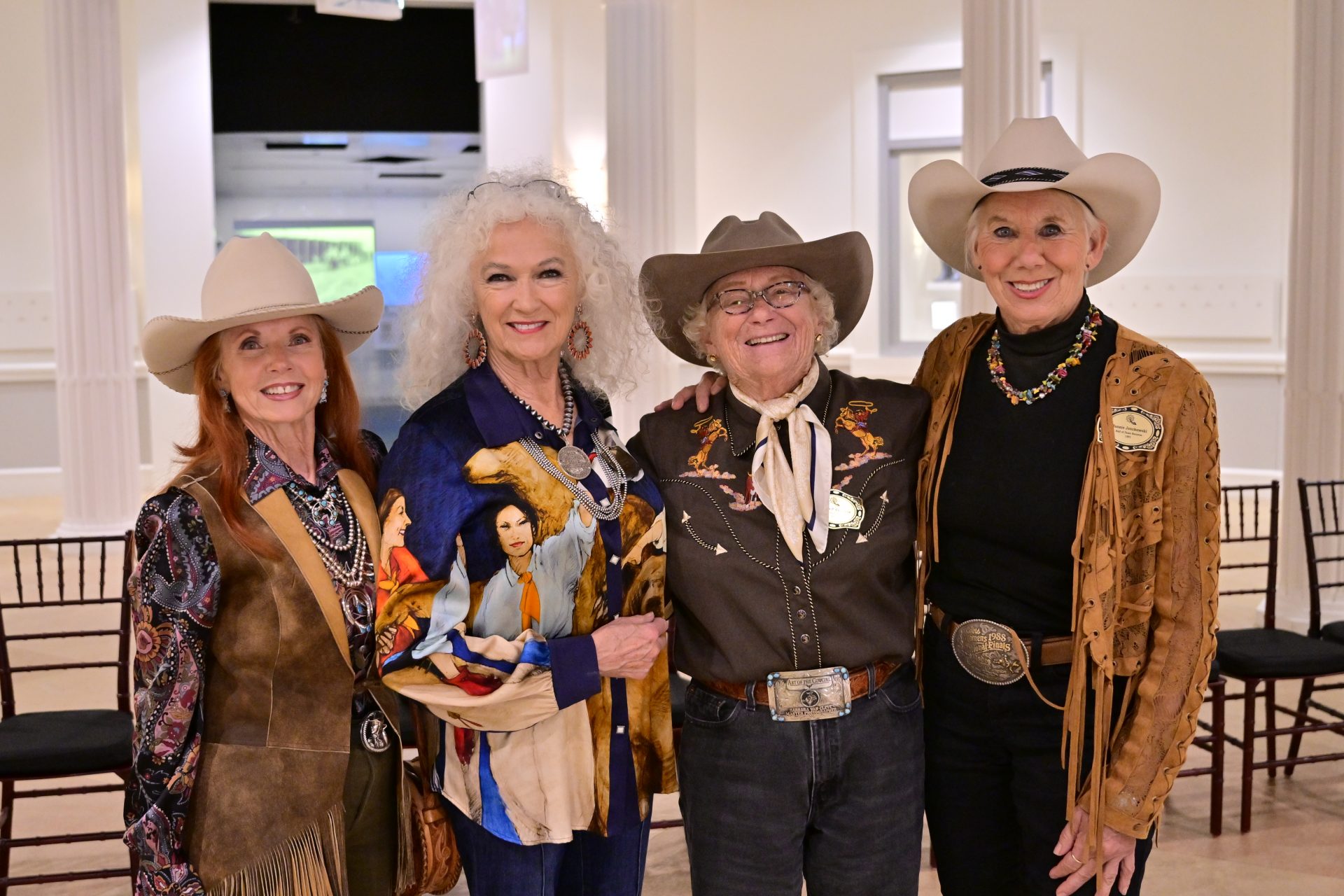 Four older women wearing cowboy hats and Western-themed outfits stand together smiling in a bright room with white walls and empty chairs in the background.