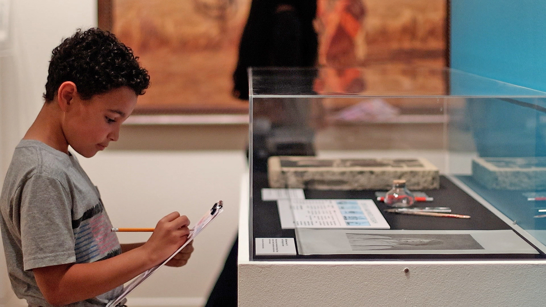 Boy in a gray t-shirt writes on a clipboard beside a museum display case with artifacts inside a glass enclosure.