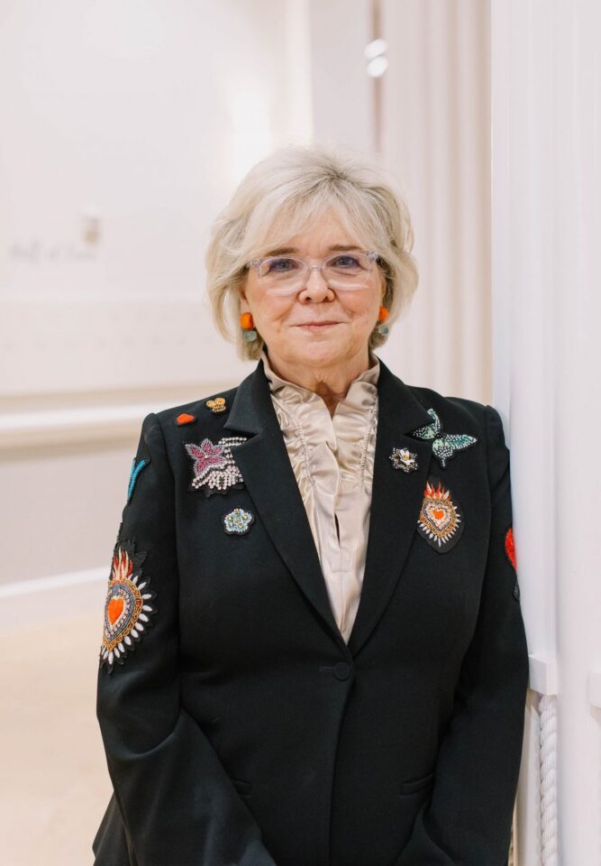 Older woman in a black blazer with embroidered patches, standing beside white vertical pillars in a light hallway.