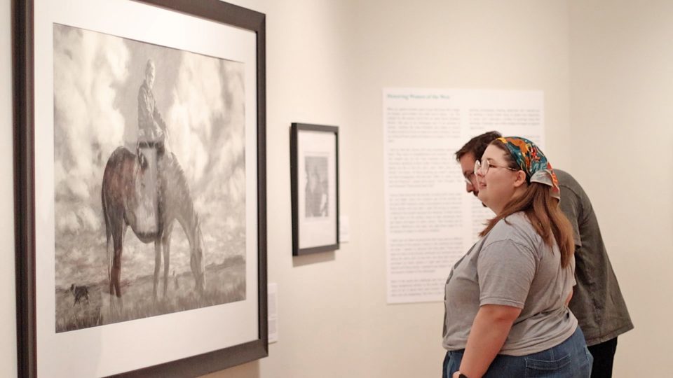 Two visitors in a gallery view a large black-and-white photo of a person riding an elephant; wall text panels are nearby.