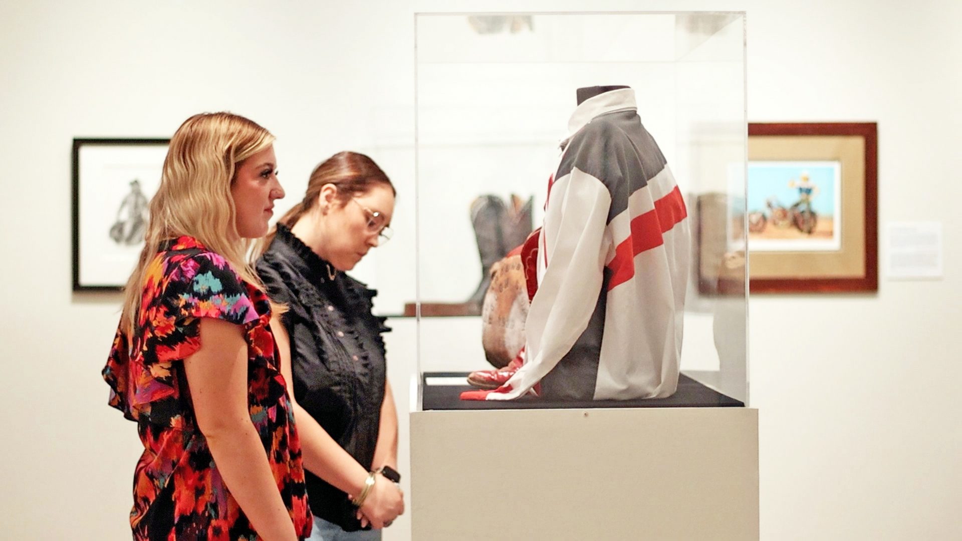 Two women observe a sports jacket and gloves displayed in a museum exhibit case. Framed artwork hangs on the white walls behind them.
