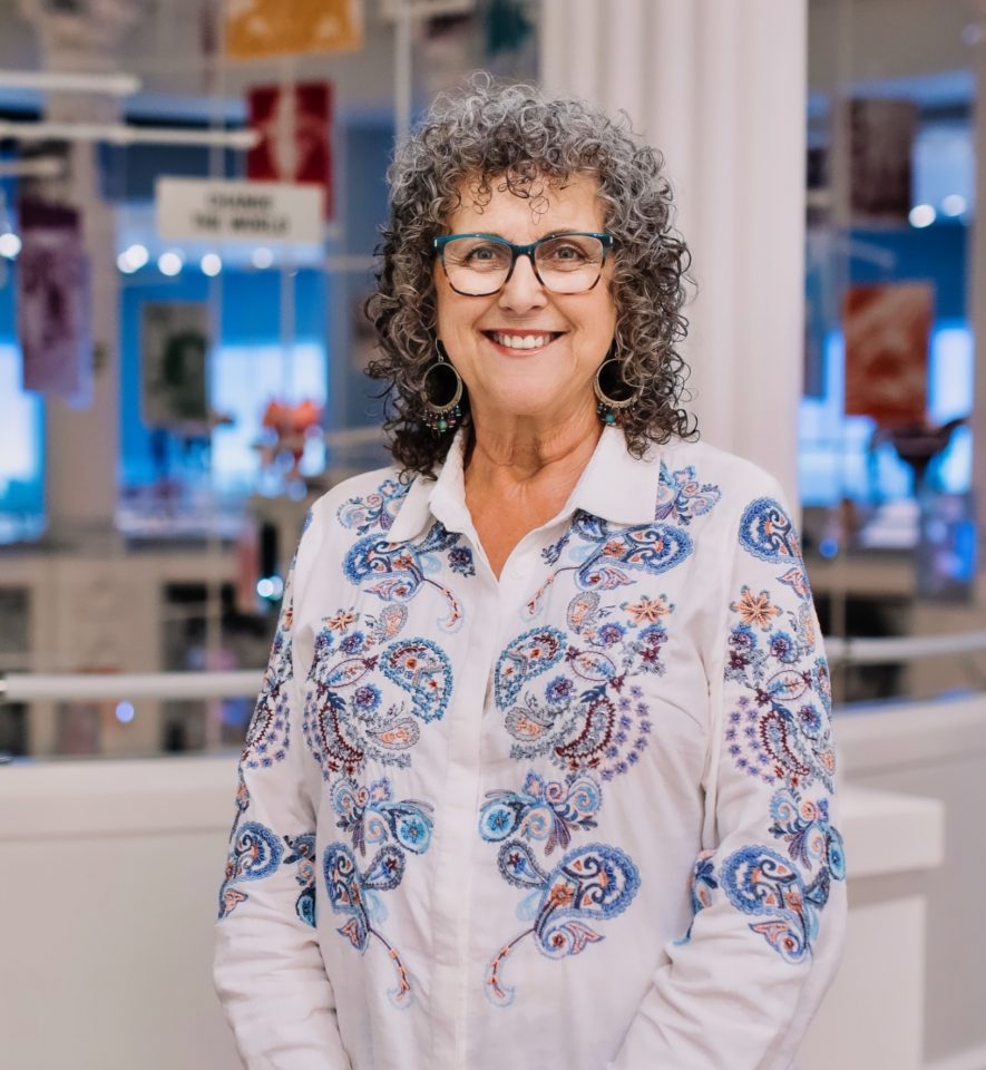 Smiling woman with curly gray hair, blue glasses, and a white embroidered blouse standing indoors.
