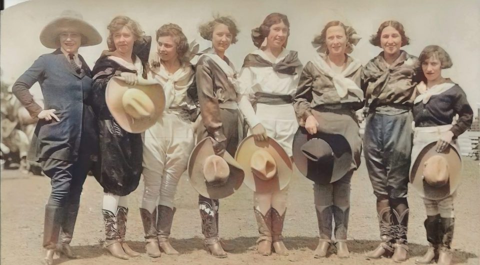 Group of nine women in vintage Western clothing posing outdoors, holding wide-brim hats and smiling together.
