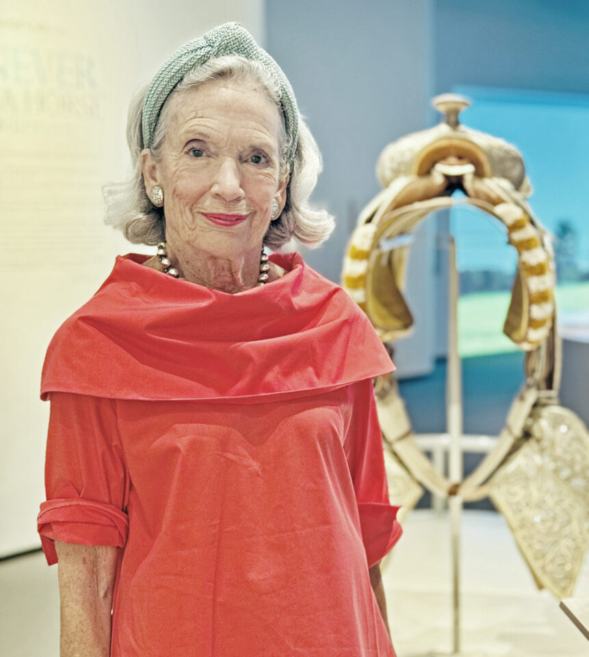 Older woman in a red dress with a green headband and pearl earrings posing near a decorative horse sculpture in a museum/gallery setting.