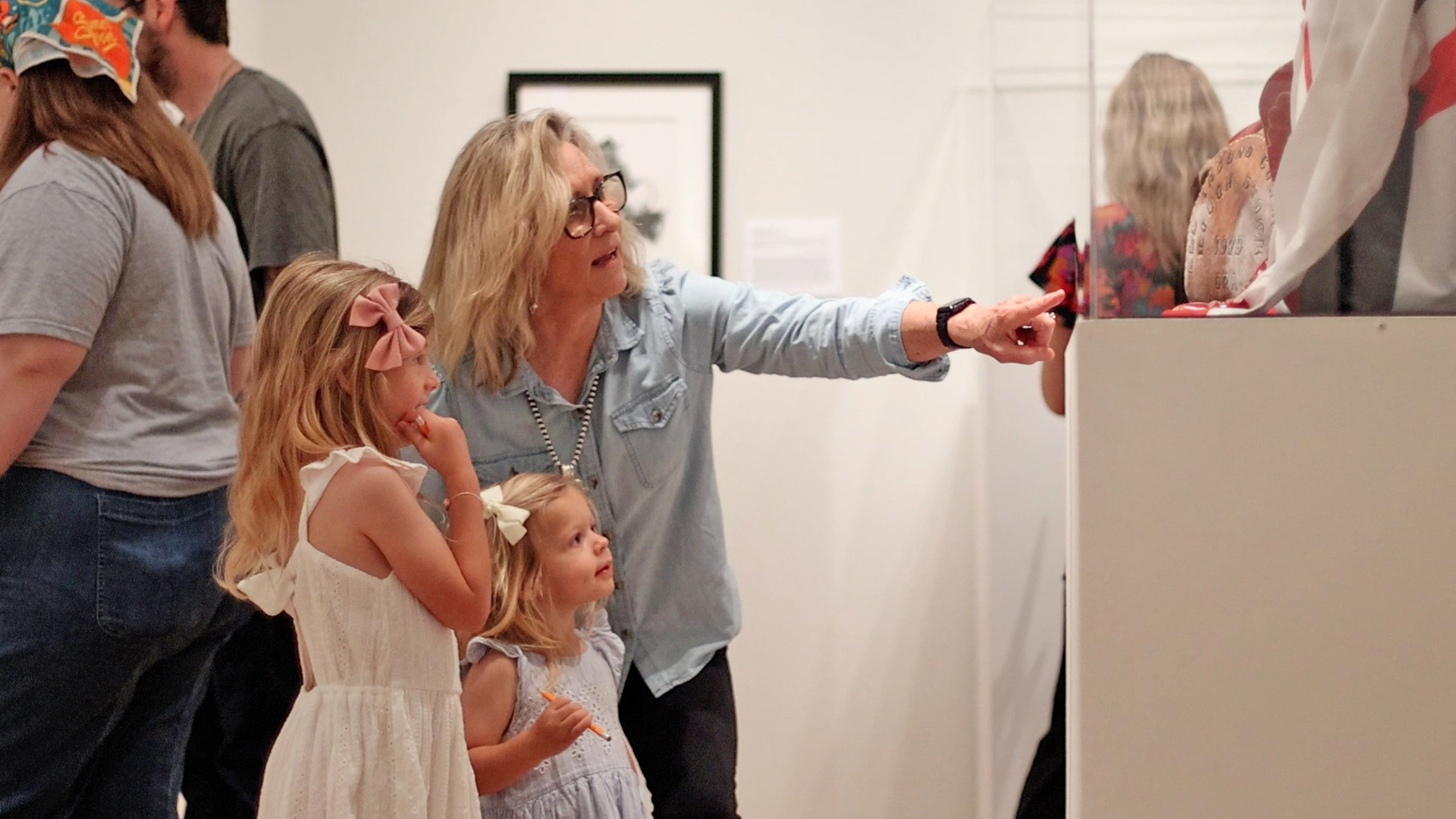 Woman in a denim shirt points toward a display as two young girls in white dresses look on in a museum setting