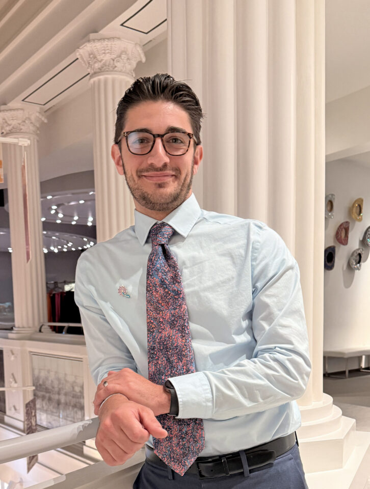 Smiling man in a light blue shirt and patterned tie leans on a railing in a bright, white showroom with decorative columns.